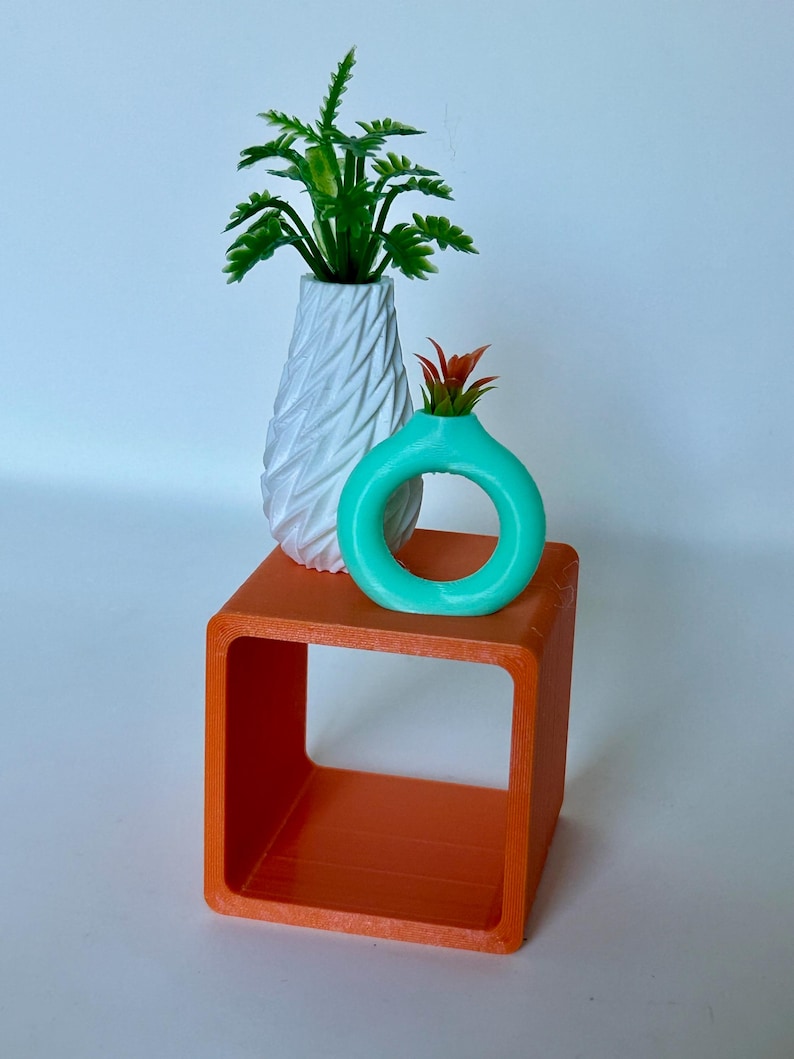 May include: Two small potted plants on a bright orange square shelf. The larger plant is in a white, textured vase. The smaller plant is in a turquoise vase shaped like a ring.