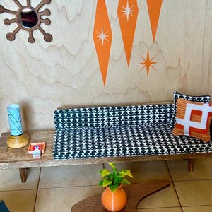 May include: A miniature mid-century modern living room set with a black and white patterned sofa, a wooden coffee table, a lamp, and a potted plant. The wall is decorated with orange diamond shapes and white stars.