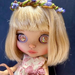May include: A doll with blonde hair and a floral crown. The doll has large eyes with glitter and freckles. She wears a pink floral dress with a matching bow and holds a basket.