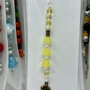 May include: A yellow beaded tassel with a flower charm. The tassel is made of small yellow beads and has a white string. The flower charm is made of metal and has a brown finish.