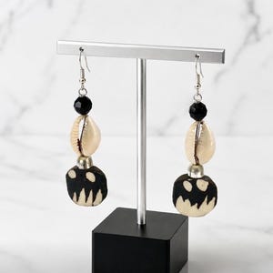 May include: A pair of dangle earrings featuring a silver-tone hook, a faceted black bead, a cream-colored cowrie shell, and a round black pendant with a jagged white pattern at the bottom, all suspended from a silver-tone metal display stand.
