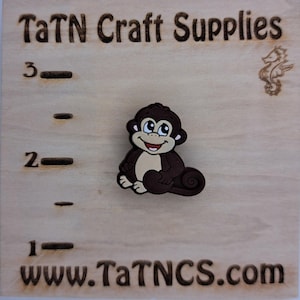 May include: A brown and tan monkey charm with a smiling face, sitting on a wooden surface. The text "TaTN Craft Supplies" and "www.TaTNCS.com" are visible. The wooden surface has measurements in inches.