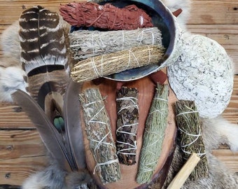 May include: An assortment of sage bundles, feathers, and a decorative shell arranged on a wooden surface. The sage bundles are various colors, including red, gray, and green, and are tied with white string. Feathers and a piece of wood are also included.