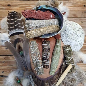 May include: An assortment of sage bundles, feathers, and a decorative shell arranged on a wooden surface. The sage bundles are various colors, including red, gray, and green, and are tied with white string. Feathers and a piece of wood are also included.