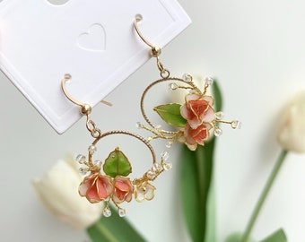 May include: A pair of gold hoop earrings with delicate floral accents. The earrings feature pink and white flowers with green leaves, and are adorned with small clear beads.