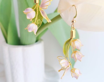 May include: A pair of delicate gold earrings featuring a cluster of pink lily of the valley flowers with green leaves. The earrings are made with wire and have a dangling design.