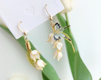 May include: A pair of gold earrings featuring a cluster of white lily of the valley flowers with a blue butterfly charm on one earring.