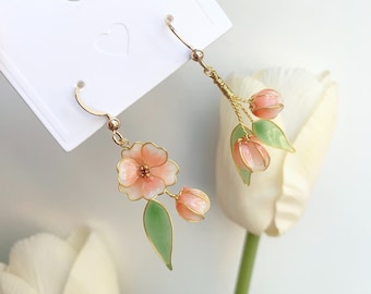 May include: Delicate floral earrings featuring peach-colored blossoms and buds with gold-tone accents. The earrings include green leaves and are attached to gold-colored ear wires. The earrings are displayed on a white card.