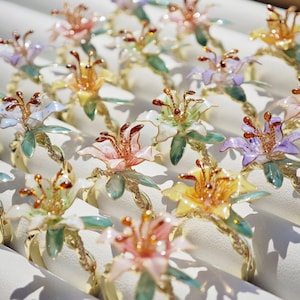 以下が含まれることがあります： ピンク、黄色、紫など、さまざまな色の花の指輪のコレクション。各リングは、金色のバンドと緑の葉が付いた詳細な花のデザインが特徴です。リングは白いディスプレイに配置されています。