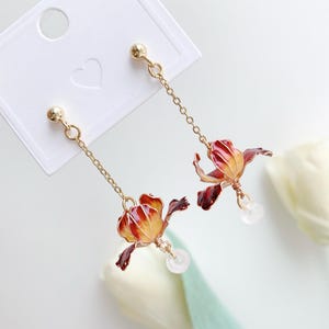 Op de afbeelding: Een paar gouden oorhangers met rode en gele bloemencharms en heldere kristallen kralen die aan de onderkant hangen.