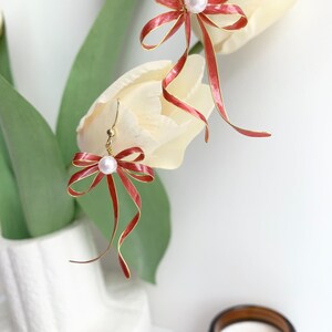 Op de afbeelding: Een paar gouden oorbellen met rode strikken van lint en witte parel accenten. De oorbellen hangen aan een witte tulp.