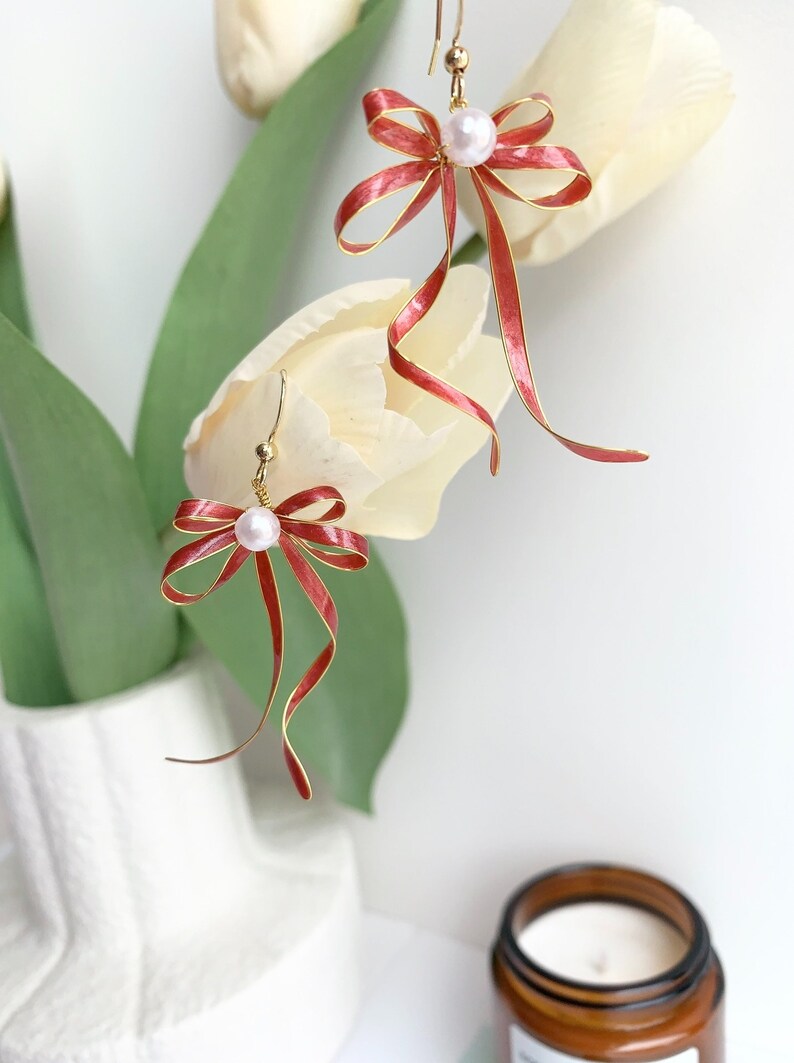 Op de afbeelding: Een paar gouden oorbellen met rode strikken van lint en witte parel accenten. De oorbellen hangen aan een witte tulp.