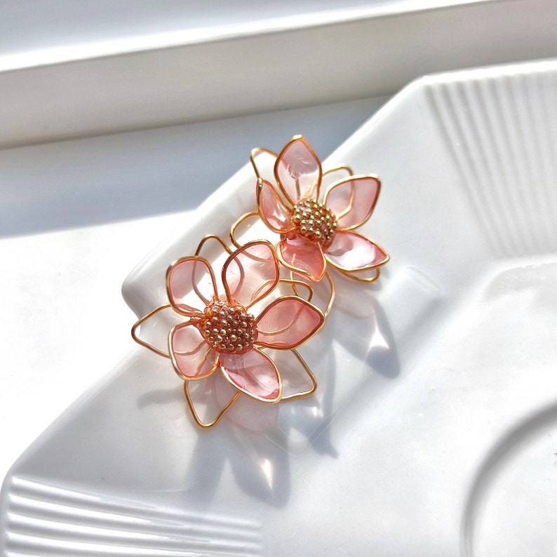 May include: A pair of pink flower-shaped earrings with gold wire accents. The earrings are made of translucent pink plastic and have a delicate, feminine design.