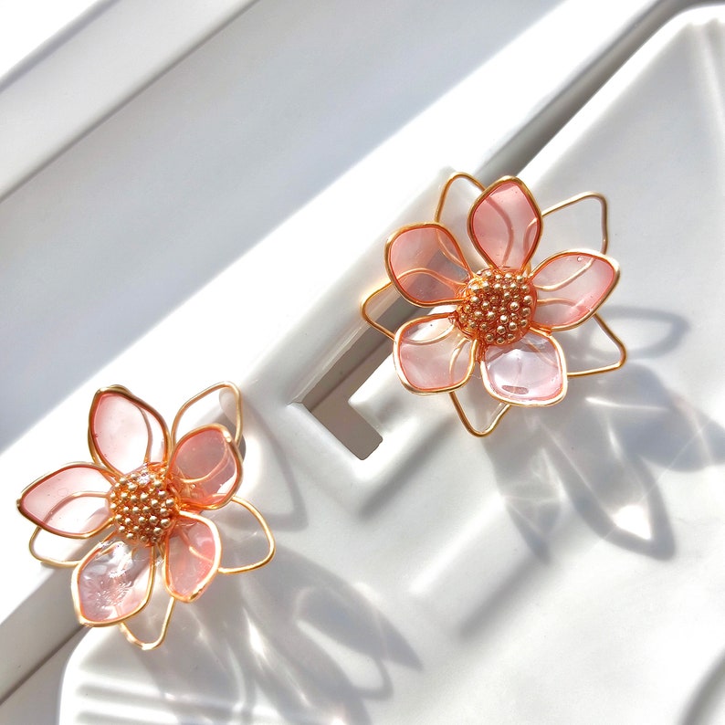 May include: A pair of gold earrings with pink flower designs. The flowers are made of translucent pink material and have a gold wire frame.