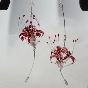 May include: A pair of silver earrings with red flower accents. The flowers are made of glass and are shaped like lilies. The earrings are hanging from a silver hook.