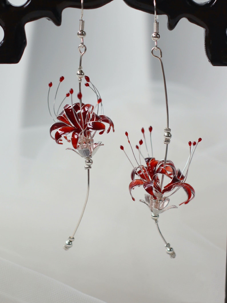 May include: A pair of silver earrings with red flower accents. The flowers are made of glass and are shaped like lilies. The earrings are hanging from a silver hook.