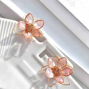 May include: A pair of delicate pink flower earrings with gold wire accents. The flowers have a layered petal design and a small cluster of gold beads in the center.