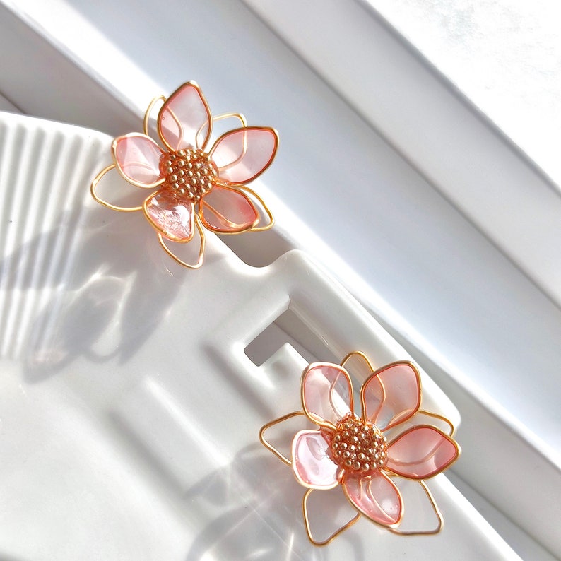 May include: A pair of delicate pink flower earrings with gold wire accents. The flowers have a layered petal design and a small cluster of gold beads in the center.