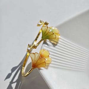 Könnte beinhalten: Ein goldfarbener Ring mit einem Ginkgoblatt-Design. Der Ring hat zwei stilisierte Ginkgoblätter in Gelb- und Orangetönen mit einer kleinen Ansammlung klarer Kristalle. Der Ring ist vor einem weißen Hintergrund platziert.