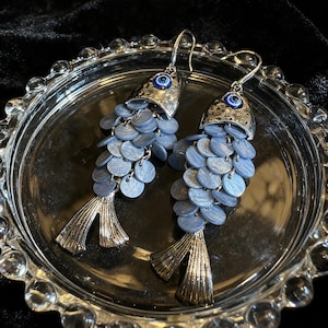 May include: A pair of fish-shaped earrings. The earrings feature a silver-colored metal head with a blue eye detail and a cascade of small, round, blue-grey discs. The tail fins are also silver-colored metal. The earrings are displayed on a silver-colored, decorative plate.