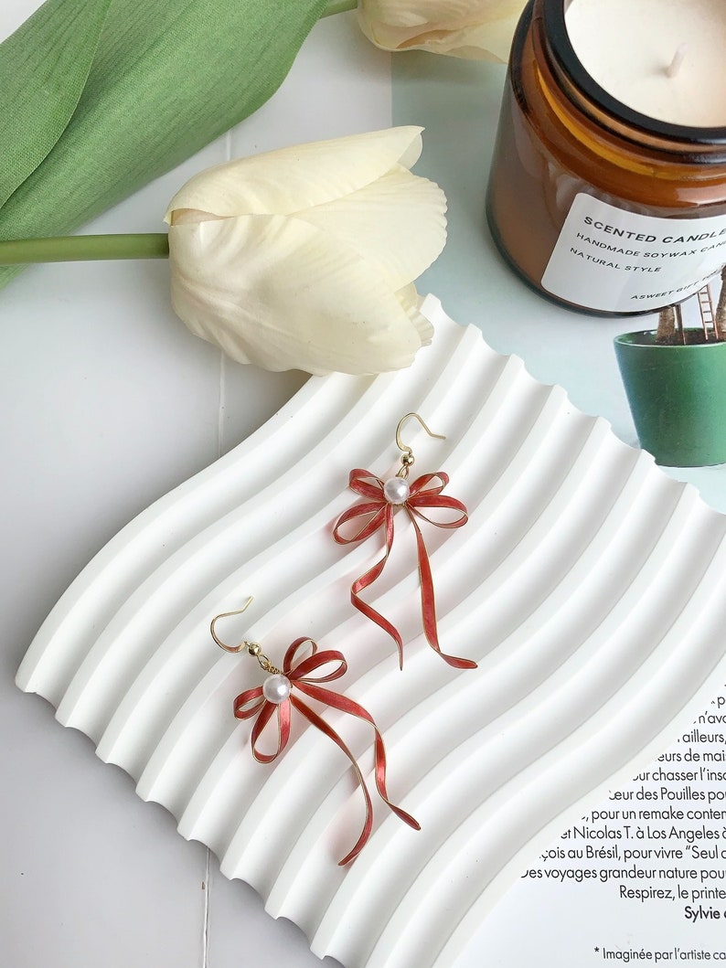 Op de afbeelding: Een paar gouden oorbellen met rode strikken en witte parel accenten. De oorbellen liggen op een wit oppervlak met een witte tulp op de achtergrond.