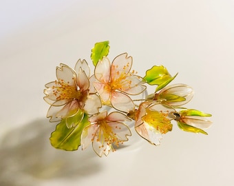 May include: A delicate cluster of translucent pink and white cherry blossoms with gold accents and green leaves. The blossoms are made of glass and are arranged in a bouquet.