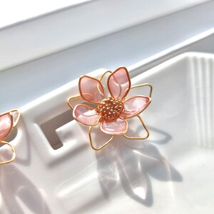 May include: A pair of pink flower earrings with gold wire accents. The flowers are made of translucent pink resin and have a delicate, feminine look.