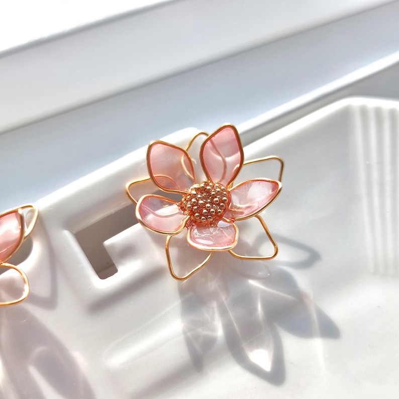 May include: A pair of pink flower earrings with gold wire accents. The flowers are made of translucent pink resin and have a delicate, feminine look.