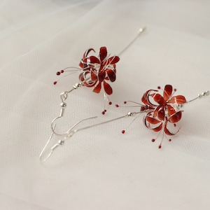 May include: A pair of silver dangle earrings with red flower-shaped charms. The flowers have a delicate, intricate design and are accented with small red beads.