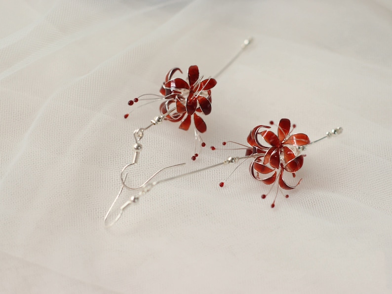 May include: A pair of silver dangle earrings with red flower-shaped charms. The flowers have a delicate, intricate design and are accented with small red beads.
