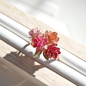 May include: A floral ring with a gold-coloured band, showcasing a cluster of flowers in pink, red, and orange hues. The ring features green leaves and a bouquet-style arrangement of the flowers.