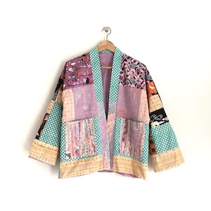 May include: A patchwork kimono-style jacket with a Halloween theme. The jacket features various fabric patterns, including polka dots, spiderwebs, and Halloween-themed prints. The color palette includes purple, teal, and beige.