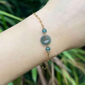 Puede incluir: Una delicada pulsera de cadena dorada que presenta un disco central circular de piedra verde similar al jade, flanqueado por dos cuentas redondas más pequeñas a juego. La pulsera está asegurada con una fina cadena de eslabones dorados.