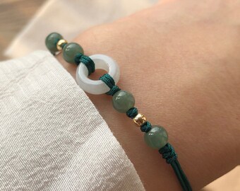 May include: A green jade bracelet with a white jade ring charm and gold beads. The bracelet is made with a dark green cord.