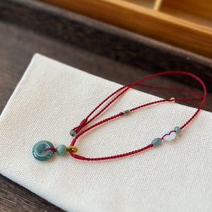 May include: A red cord bracelet featuring a circular green jade donut pendant, a small green jade bead, and a yellow loop connector. The bracelet includes additional small green and white beads, a metallic silver accent bead, and an adjustable knot closure.