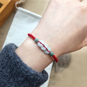 May include: A red string bracelet with three white jade beads. The beads are round and have a slightly green hue.