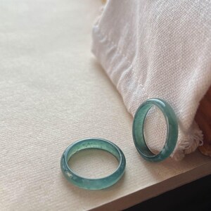 May include: Two jade green rings, one slightly larger than the other, are displayed on a white surface. The rings are smooth and have a subtle shimmer.