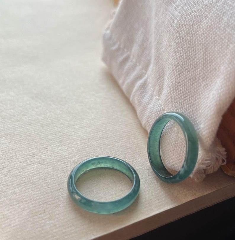 May include: Two jade green rings, one slightly larger than the other, are displayed on a white surface. The rings are smooth and have a subtle shimmer.