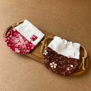 Puede incluir: Dos pequeñas bolsas con cordón que presentan una parte superior de tela blanca y una base estampada. Una bolsa tiene una base roja con flores de cerezo rosas, mientras que la otra tiene una base marrón oscuro con flores blancas y una perla.