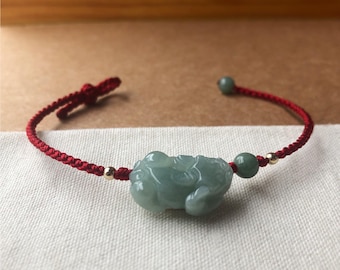 May include: A bracelet featuring a central carved light green jade animal charm on a thin red braided cord. The design includes two small gold-toned metallic beads and one additional round light green jade bead, with adjustable knotted ends.