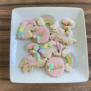 May include: A set of unicorn-themed sugar cookies decorated with pastel colors and gold accents. The cookies feature a unicorn, a rainbow, and the word "three".