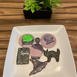 May include: A white square plate displays Halloween-themed sugar cookies. The cookies include a cauldron with green bubbling potion, a potion bottle, a crystal ball, a black cat, a witch hat, and a spell book with the word "Spells".