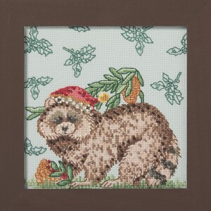 May include: A framed cross-stitch embroidery featuring a brown raccoon wearing a red Santa hat with white trim. The raccoon is positioned against a light blue background patterned with green holly leaves. A small orange fruit sits near the raccoon.