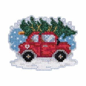May include: A cross-stitch ornament featuring a red car carrying a green Christmas tree on its roof. The car has black wheels and is set against a snowy, blue background. A heart-shaped detail is on the car's front.