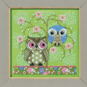 May include: Two owls sitting on a branch in a green and pink floral pattern. The owl on the left is brown with pink eyes and the owl on the right is blue with black eyes.