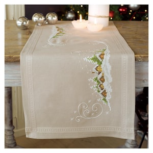 Könnte beinhalten: Beiger Weihnachtstischläufer mit gestickter Winterszene. Das Design zeigt grüne und goldene Bäume, weißen Schnee und dekorative Wirbel. Der Läufer liegt auf einem Holztisch mit Kerzen und Ornamenten.
