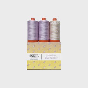 Puede incluir: Tres carretes de hilo en tonos morado claro y crema, cada uno con una base naranja, se muestran sobre una caja amarilla. La caja presenta un patrón repetitivo de pétalos morados y el texto Aurifil Color Builder Hawaiian Blue Ginger.