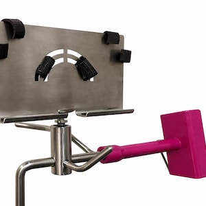 May include: A stainless steel stand featuring a rectangular plate with black straps and a curved cutout, mounted on a central pole. A bright pink, rectangular handle is attached to the side of the metal assembly via a horizontal arm.