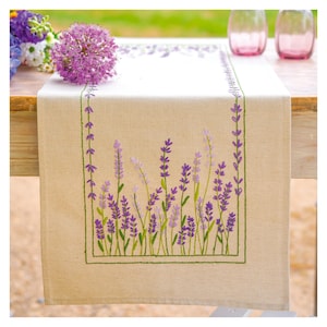 May include: A beige linen table runner with embroidered lavender sprigs in shades of purple and green. The runner features a rectangular design with a border of lavender and a central field of lavender stems. The runner is displayed on a wooden table.