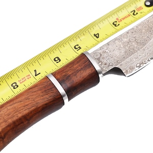 May include: A brown wooden handled knife with a silver blade. The blade has a patterned finish. The knife is measured against a yellow tape measure, showing it is 8 inches long.